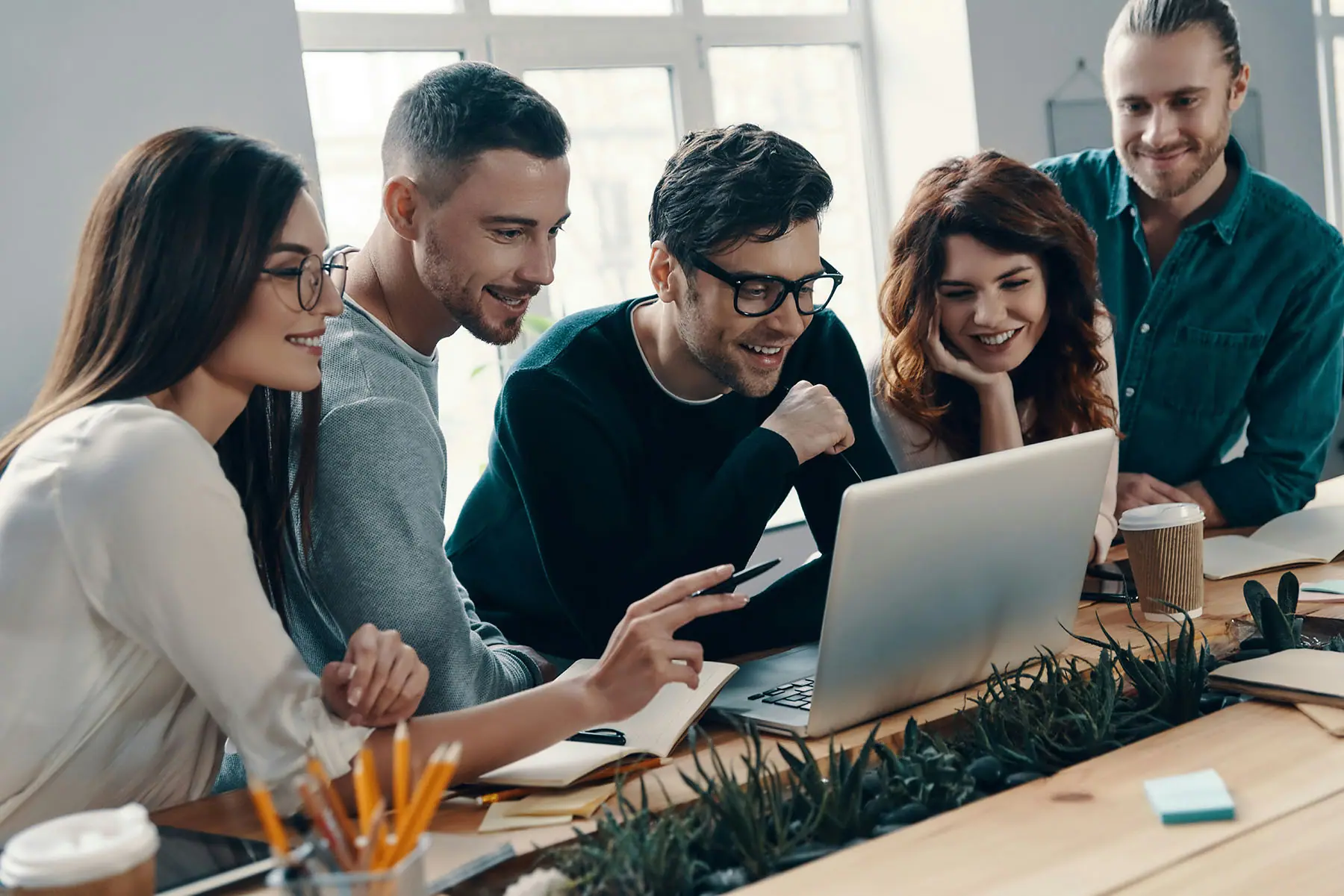 Five colleagues gathered around a laptop at a shared desk, working through something together